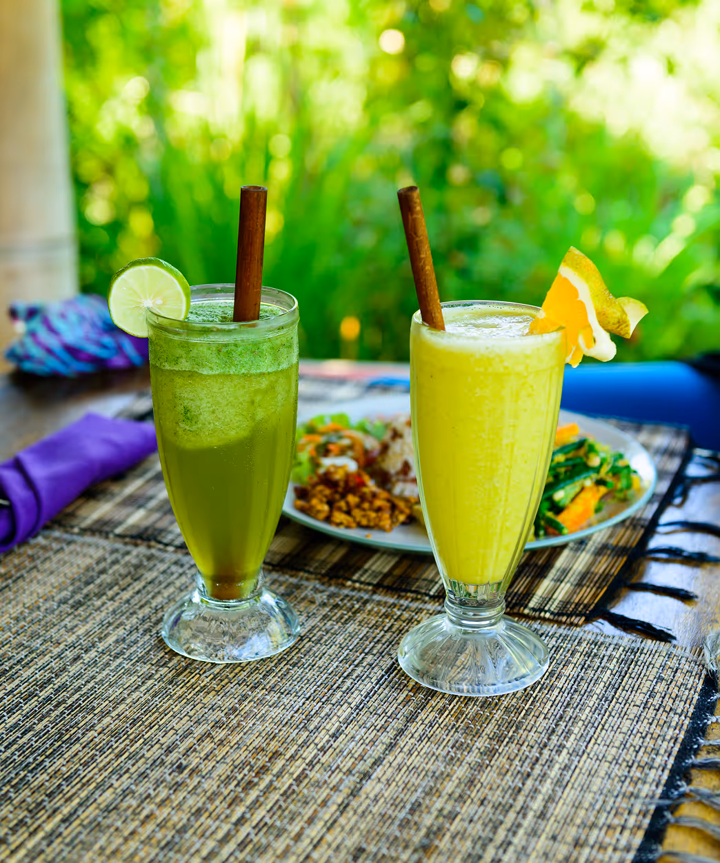 Two tall glasses of fresh green and yellow juice with wooden straws, garnished with lime and orange slices, on a woven placemat with a plate of food in the background.